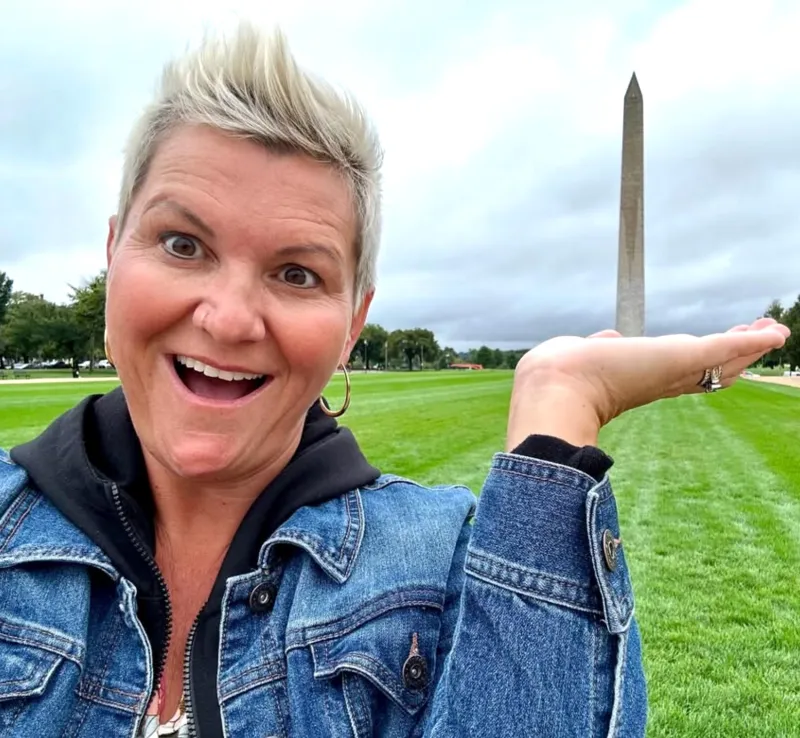 Shelly Shoultes in a denim jacket, delighted, holding her hand out so the Washington Monument looks like it is standing on her palm